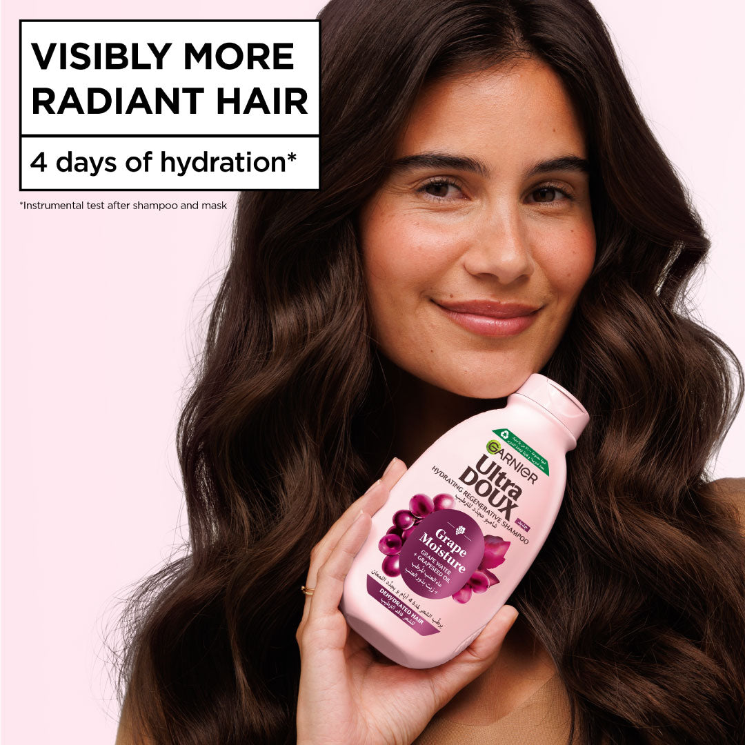 Woman with long, wavy hair holding a bottle of Garnier Ultra Doux shampoo against a pink background.